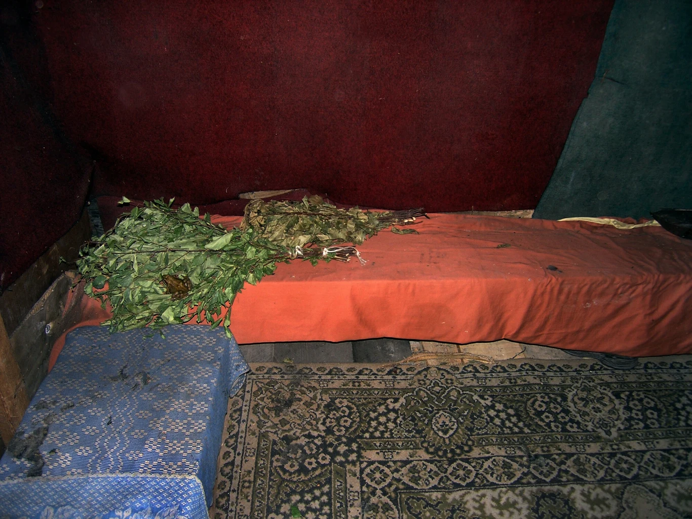 Łóżko z czerwonym prześcieradłem, na którym suszy się marihuana, obok mały stolik przykryty niebieskim obrusem, całość na tle czerwonej ściany i wzorzystego dywanu.