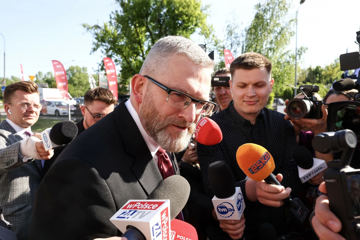 Grzegorz Braun zajął stanowisko przed drugą turą wyborów prezydenckich Mężczyzna w garniturze otoczony dziennikarzami z mikrofonami różnych stacji telewizyjnych na konferencji prasowej na zewnątrz. W tle widać drzewa i flagi reklamowe.