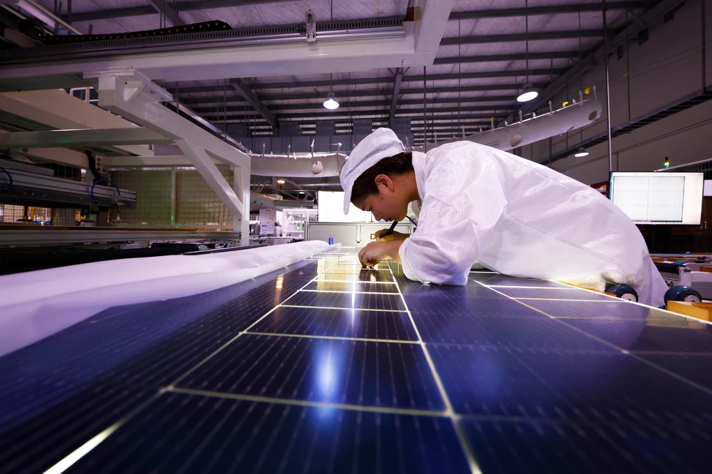 Zamknięcie się na fotowoltaikę z Azji może wyhamować transformację energetyczną - obawiają się eksperci Pracownik w białym kombinezonie przeprowadza kontrolę panelu słonecznego w nowoczesnym zakładzie produkcyjnym. Metalowa konstrukcja hali, oświetlenie jarzeniowe i precyzyjna praca nad technologią odnawialną są widoczne na pierwszym planie.