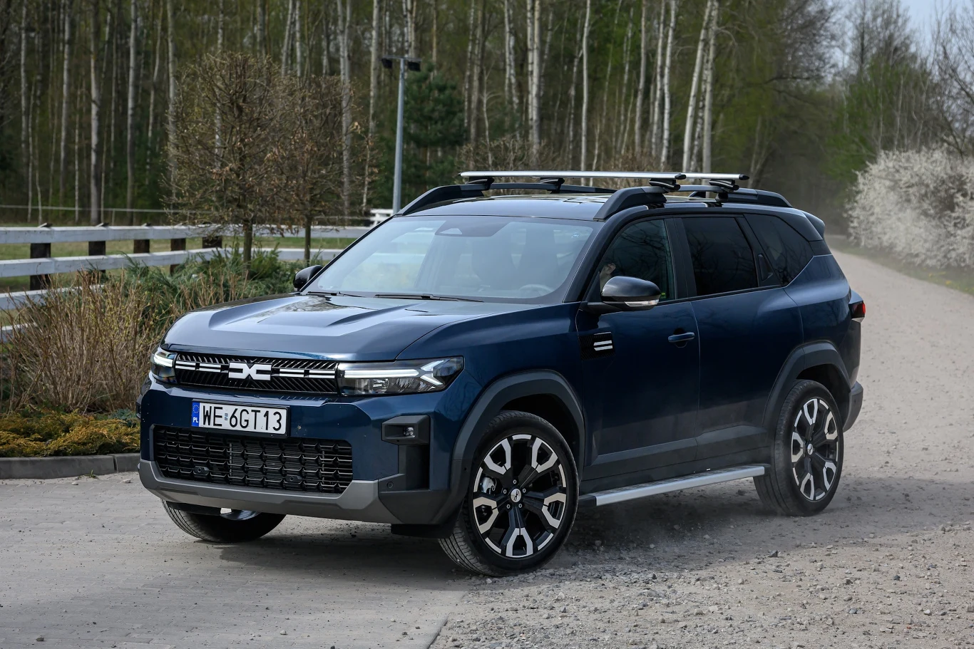 Samochód terenowy na wiejskiej drodze, otoczony przez drzewa. Nowoczesne, niebieskie auto z relingami dachowymi i charakterystyczną kratką z przodu. Tło stanowi jasnozielony las i ogrodzenie po lewej stronie.