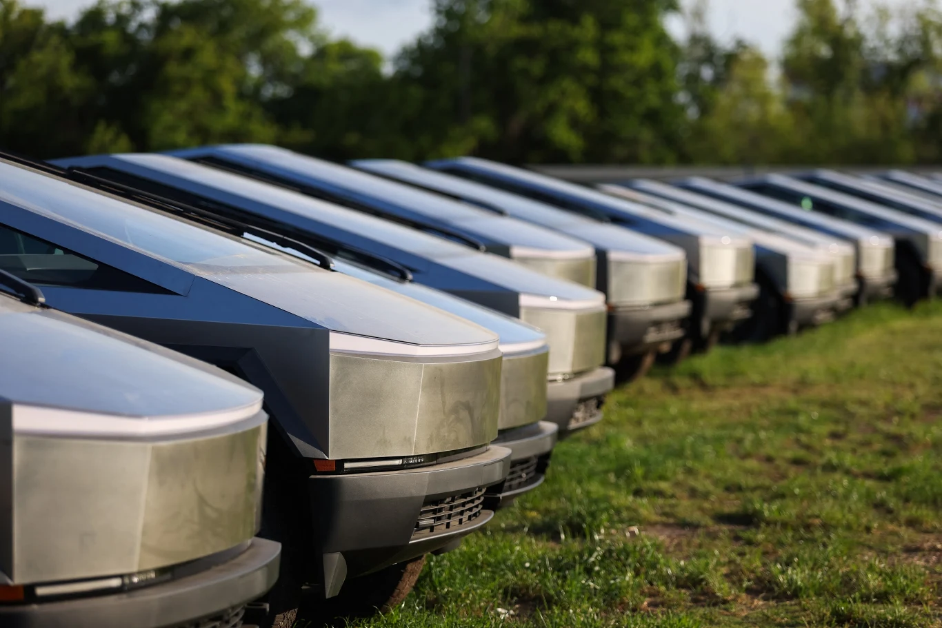 Rząd nowoczesnych samochodów o futurystycznym wyglądzie, ustawionych na trawiastej powierzchni, otoczonych zielenią drzew w tle. Metaliczna powłoka i unikalny kształt pojazdów przyciągają uwagę.