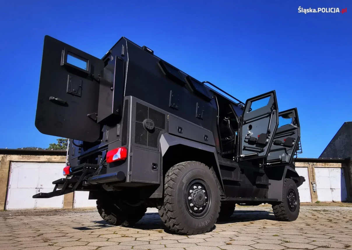 Czarny pojazd opancerzony stoi na placu betonowym, z otwartymi drzwiami po prawej stronie. Pojazd charakteryzuje się dużymi kołami terenowymi oraz solidną konstrukcją, co podkreśla jego funkcję w siłach porządkowych.