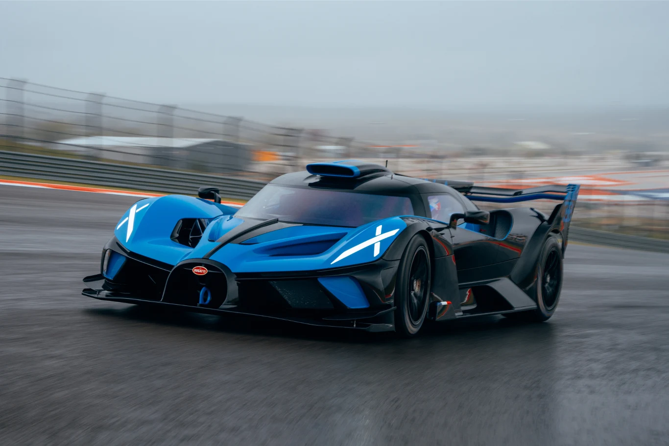 Wyścigowy samochód sportowy w kolorze niebieskim z czarnymi elementami, porusza się z dużą prędkością na mokrej nawierzchni toru wyścigowego. Nowoczesne i aerodynamiczne kształty auta podkreślają jego szybki charakter.