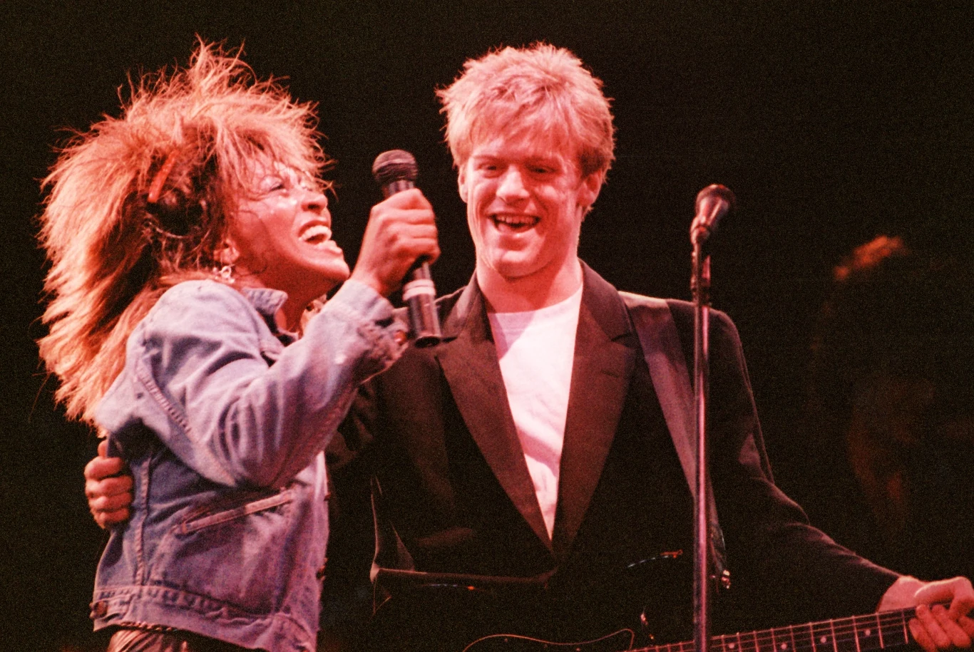 Tina Turner i Bryan Adams w 1985 r. Dwie osoby, mężczyzna z gitarą i kobieta w dżinsowej kurtce, śpiewające razem na scenie w trakcie występu muzycznego. Oboje są uśmiechnięci i wyglądają na zaangażowanych w występ.