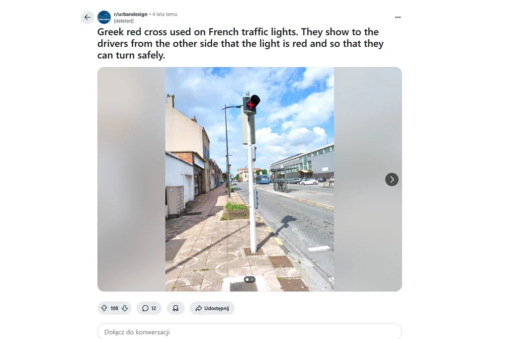 Sygnalizator z czerwonym plusem, lub greckim krzyżem, pełni istotną funkcję informacyjną / Fot. urbandesign@reddit Sygnalizacja świetlna na słupie przy ulicy, z czerwonym krzyżem widocznym na górze słupa. W tle widać chodnik i fragmenty budynków. Jasne niebo z lekkim zachmurzeniem dopełnia sceny.