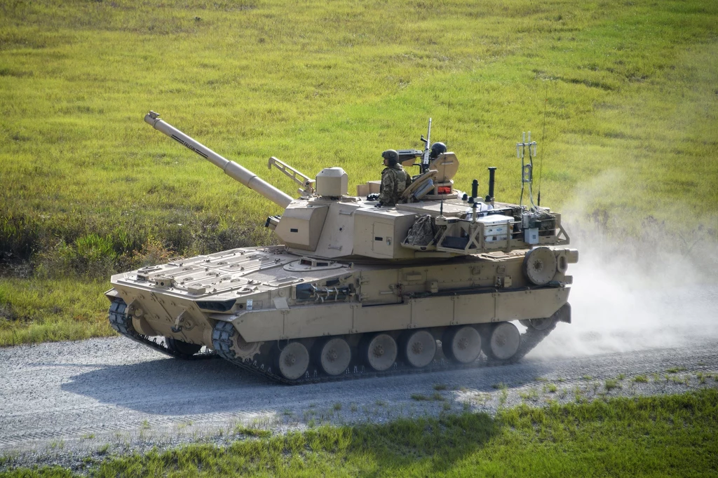 M10 Booker jest często nazywany "lekkim czołgiem", jednak U.S. Army nie klasyfikuje tak tego pojazdu Czołg porusza się po szerokiej drodze, otoczonej zielonymi polami. Widać unoszący się za nim kurz, co sugeruje dynamikę ruchu.