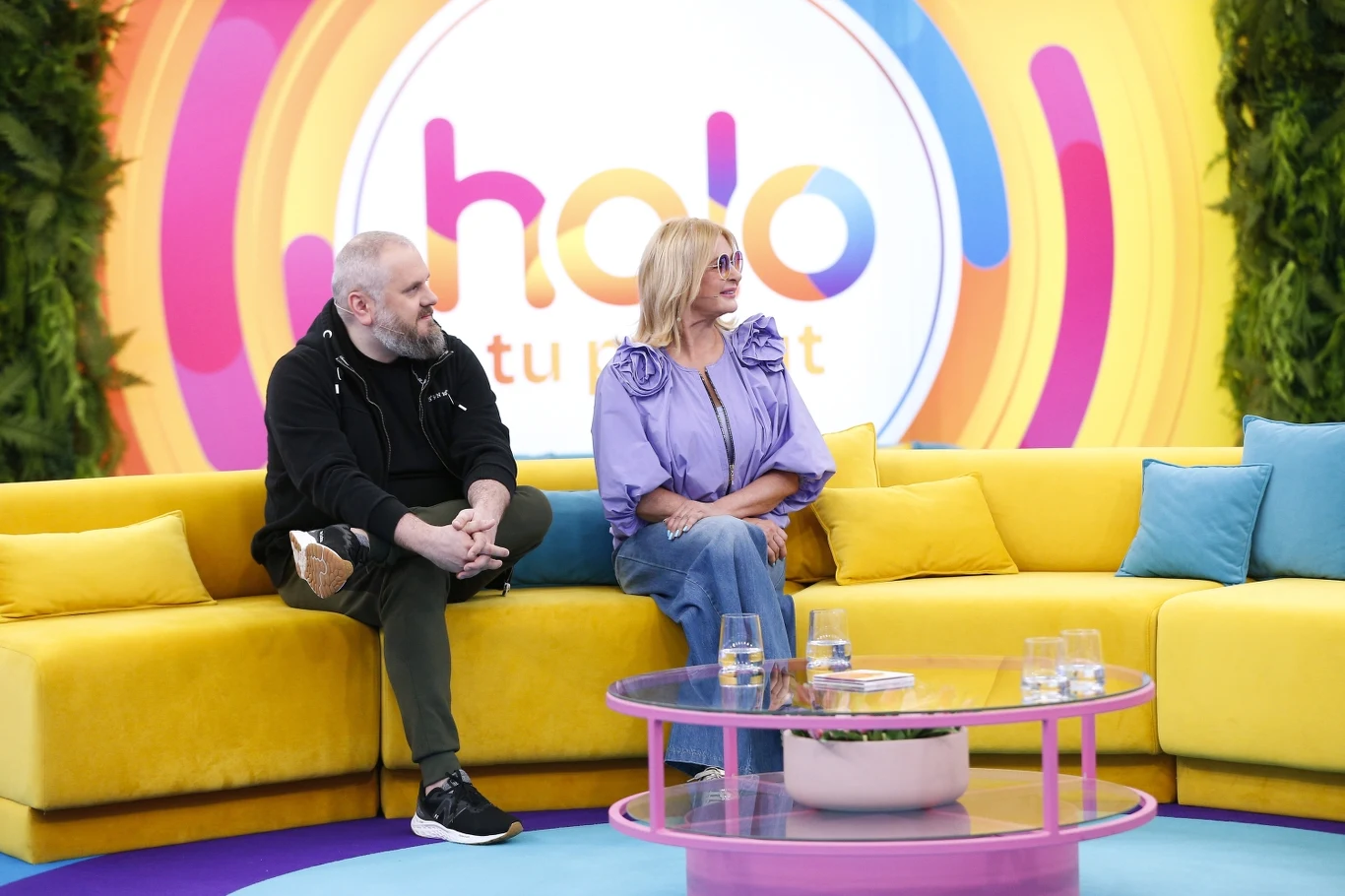 Majka Jeżowska ze swoim synem, Wojciechem Dwoje ludzi siedzi na kolorowej kanapie w studiu telewizyjnym z logiem 'halo tu dzieje' w tle, otoczonych nowoczesnym wystrojem. Przed nimi znajduje się stolik kawowy z szklankami i dekoracjami.