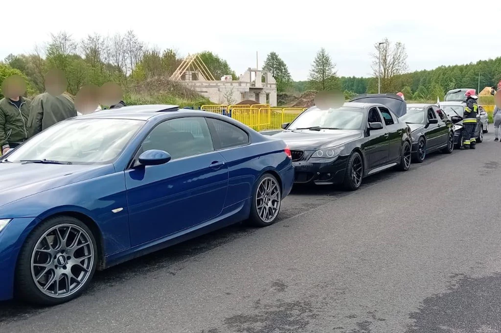 Kolizja czterech samochodów BMW / Fot: KM PSP w Toruniu Kolejka samochodów osobowych wzdłuż drogi, w tym pierwszy niebieski samochód sportowy, za którym stoją czarne pojazdy. W oddali widoczne jest budowa budynku oraz drzewa. Pojazdy i osoby na poboczu drogi.