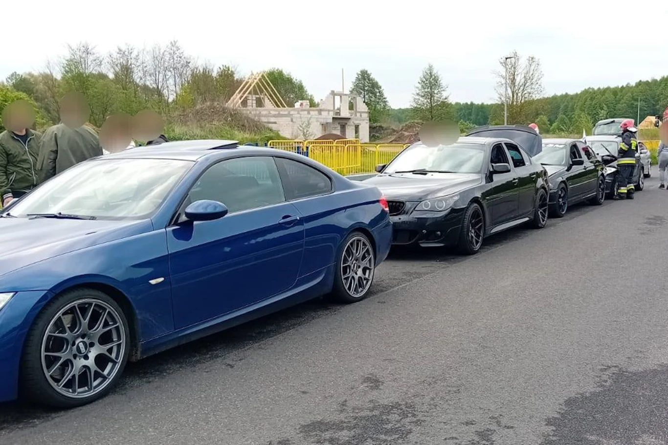 Kolejka samochodów osobowych wzdłuż drogi, w tym pierwszy niebieski samochód sportowy, za którym stoją czarne pojazdy. W oddali widoczne jest budowa budynku oraz drzewa. Pojazdy i osoby na poboczu drogi.