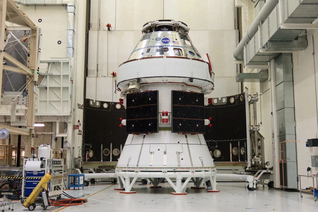 Kapsuła załogowa Orion, która zabierze astronautów NASA w misji Artemis 2 na Księżyc. Zaawansowana kapsuła kosmiczna umieszczona w hangarze technologicznym z widocznymi panelami słonecznymi i strukturami wspierającymi.