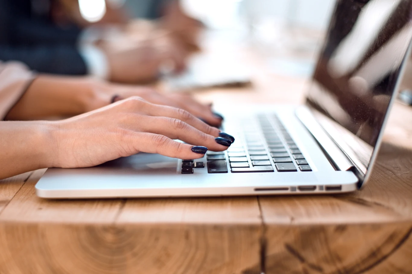 Dłonie kobiety o ciemnych paznokciach piszące na klawiaturze laptopa, urządzenie stoi na drewnianym stole w jasnym pomieszczeniu.