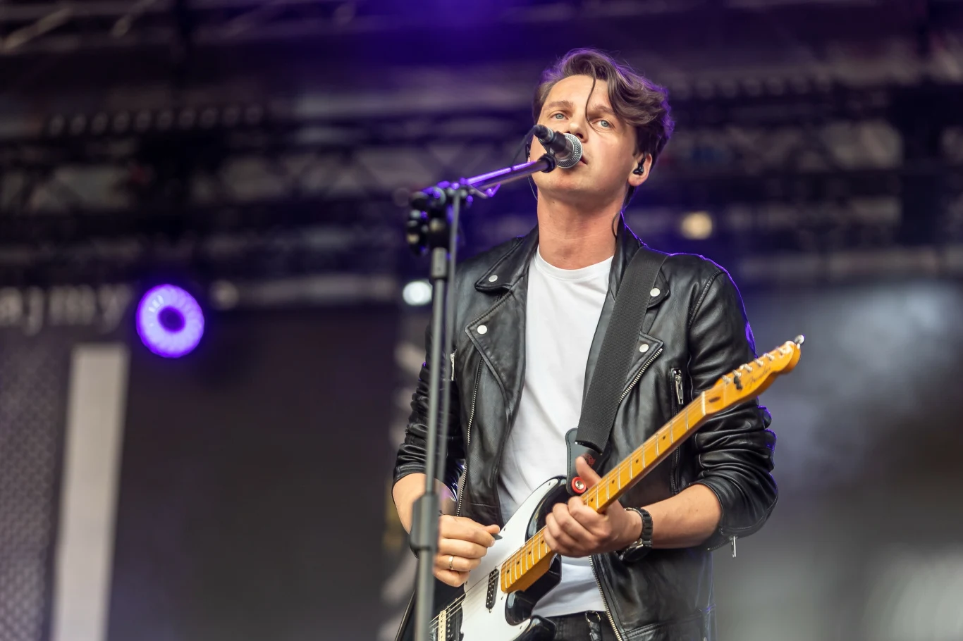 Młody mężczyzna na scenie gra na gitarze elektrycznej i śpiewa do mikrofonu. Ubrany jest w czarną skórzaną kurtkę i białą koszulkę. W tle widać sprzęt nagłaśniający i oświetlenie koncertowe.