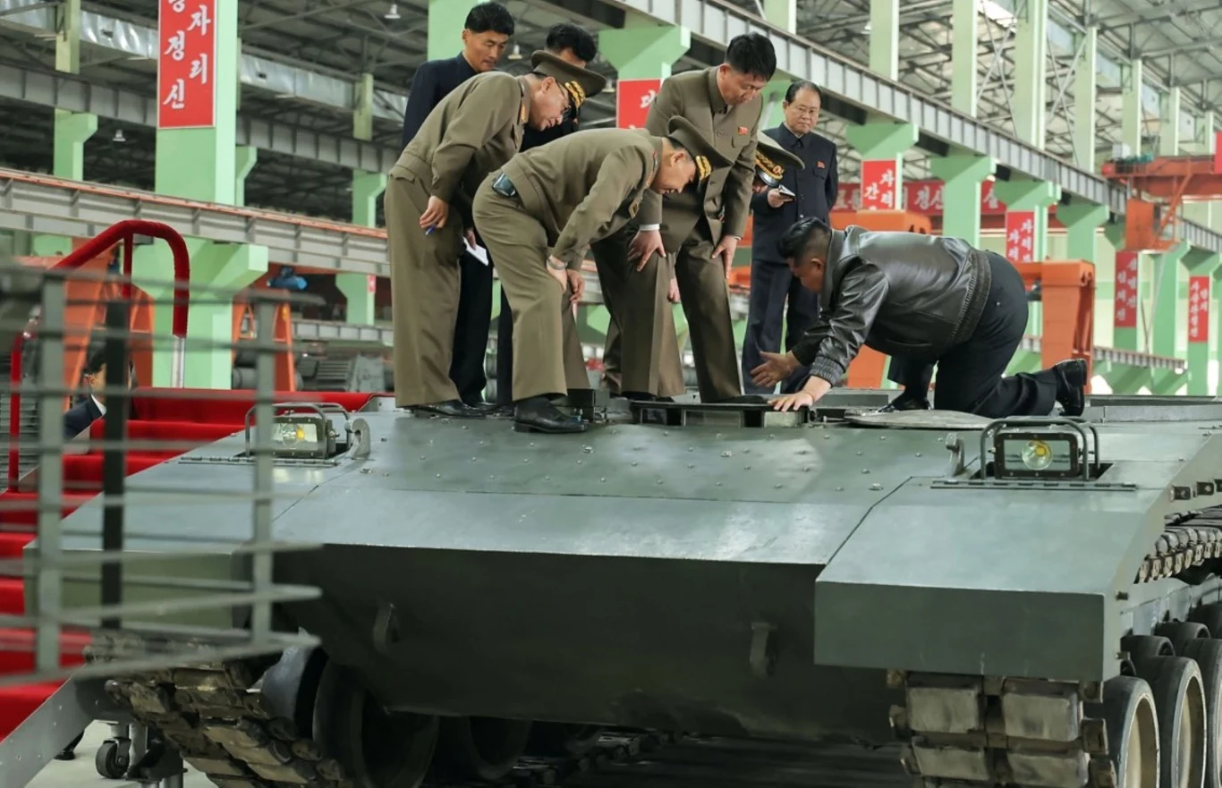 Kim Dzong Un dokonuje przeglądu czołgu rodzimej produkcji Grupa mężczyzn w uniformach wojskowych przygląda się pojazdowi opancerzonemu wewnątrz hangaru, prawdopodobnie w trakcie inspekcji lub oceny technicznej pojazdu.
