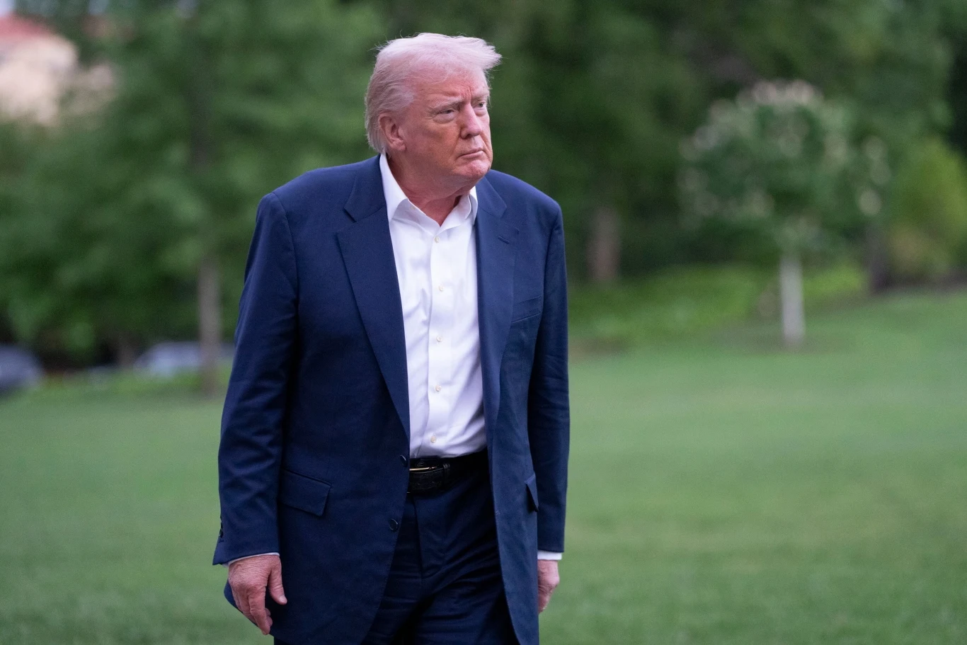 Prezydent USA Donald Trump Mężczyzna w granatowym garniturze i białej koszuli idzie po trawie na tle zieleni. Ma poważny wyraz twarzy.