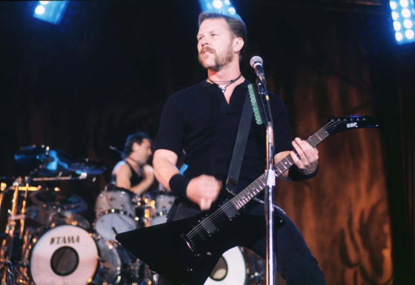 Metallica w 1996 roku Mężczyzna z czarną gitarą elektryczną, śpiewający przy mikrofonie. W tle perkusista. Oświetlenie sceniczne podkreśla atmosferę koncertu rockowego.