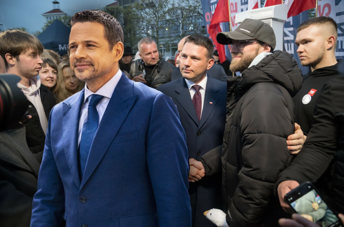 Trzaskowski wypadł najlepiej w debatach po zliczeniu wszystkich odpowiedzi w sondażu Grupa ludzi na ulicy, w tym mężczyzna w niebieskim garniturze, otoczony przez innych uczestników w kurtkach i garniturach. Na tyle widoczne są flagi i budynek.
