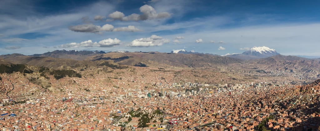 Wulkan Uturuncu widziany z La Paz w Boliwii Panorama miasta La Paz z widokiem na góry w tle. Rozległe miasto zajmuje większość dolnej części zdjęcia, a w oddali widać ośnieżone szczyty górskie. Niebieskie niebo z nielicznymi chmurami tworzy spokojne tło.