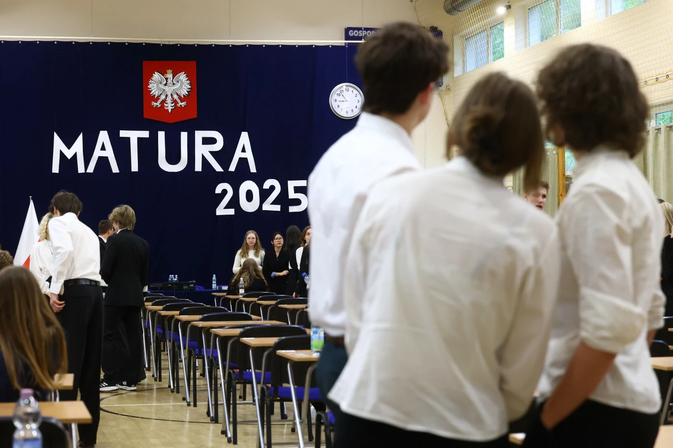 Matura 2025. Wyciekł arkusz. Mamy komentarz CKE Sala egzaminacyjna z rozstawionymi ławkami i krzesłami, na tle plakatu z napisem 'Matura 2025'. Uczniowie w eleganckich ubraniach przygotowują się do egzaminu, panuje atmosfera skupienia i oczekiwania.