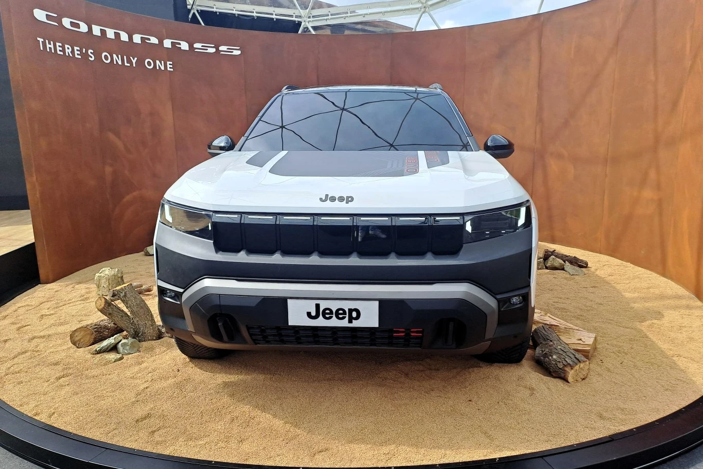 Samochód Jeep Compass ustawiony na piaskowym podłożu, z widocznym logo marki na masce i rejestracji. W tle znajduje się napis 'COMPASS' oraz hasło reklamowe 'THERE'S ONLY ONE', co wskazuje na charakter promocyjny.