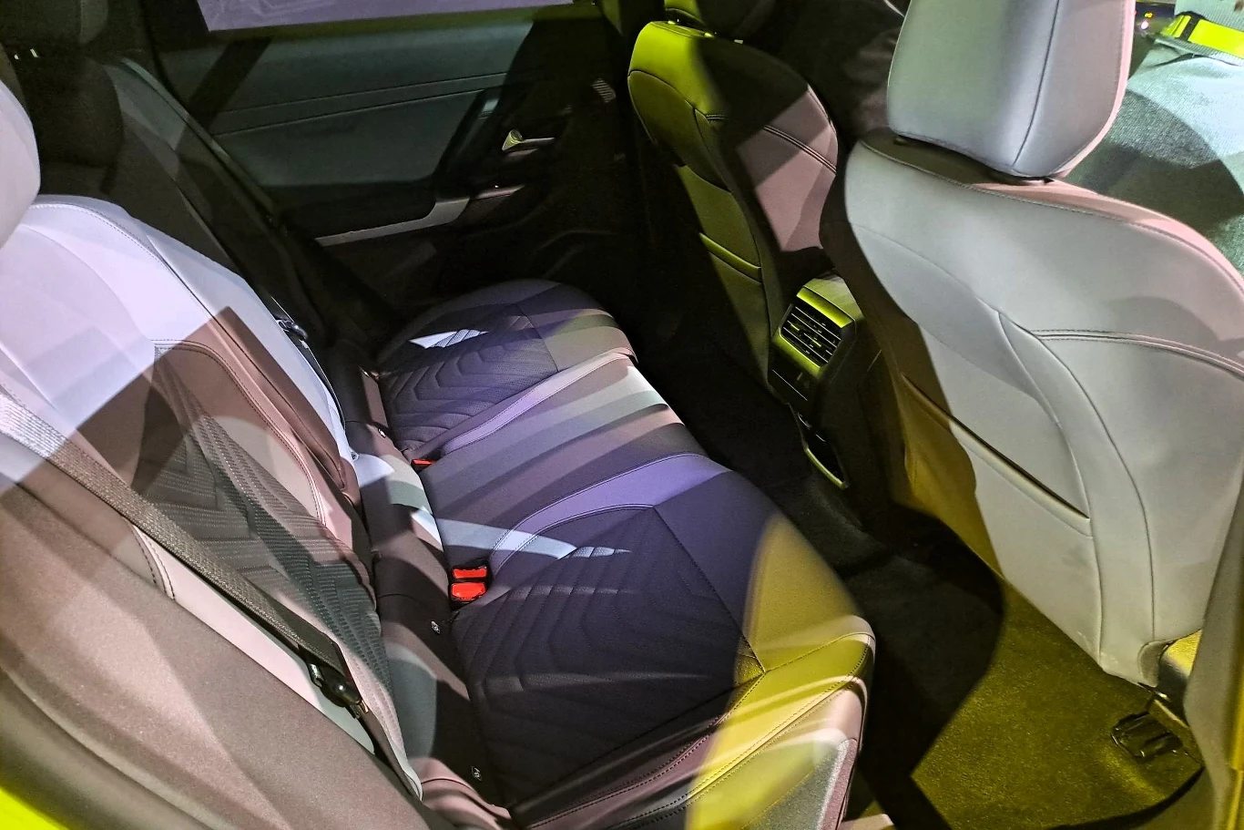 Wnętrze samochodu, tylna kanapa z trzema miejscami siedzącymi, dobrze oświetlone. Tapicerka ma nowoczesny wzór, a siedzenia są przestronne i wygodne. Widać także uchwyty na pasy bezpieczeństwa.