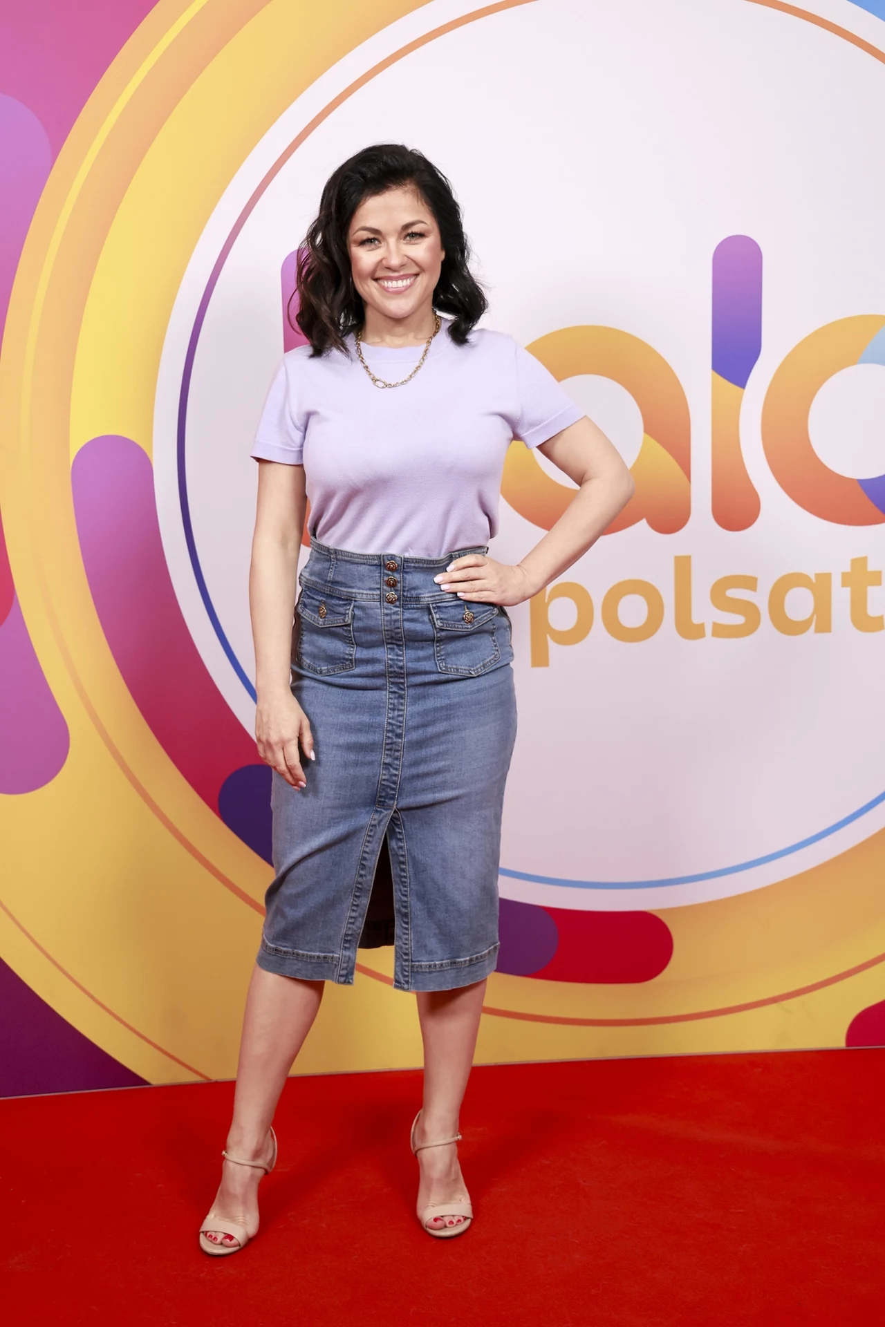 Katarzyna Cichopek zachwyciła niewymuszoną stylizacją, która może być inspiracją dla wielu kobiet Kobieta w fioletowej bluzce i dżinsowej spódnicy stoi na czerwonym dywanie przed logotypem Polsatu. Uśmiecha się do kamery.