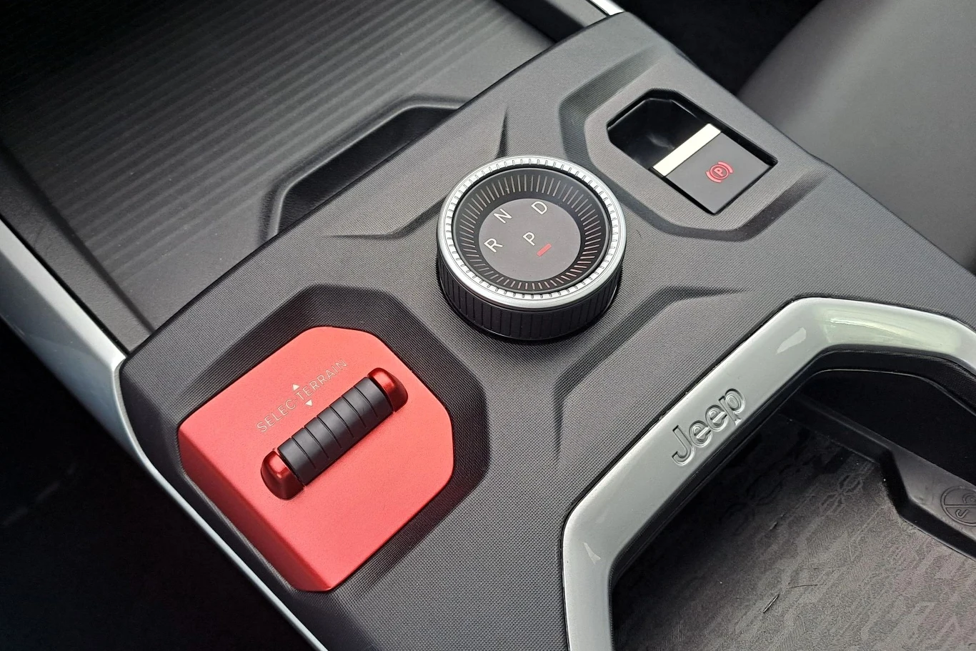 Panel sterowania w samochodzie Jeep, zawierający pokrętło zmiany biegów z oznaczeniami P, R, N, D oraz przycisk elektronicznego hamulca ręcznego. Elementy utrzymane w kontrastujących kolorach czerwonym i czarnym, z wyraźnym logo Jeep.