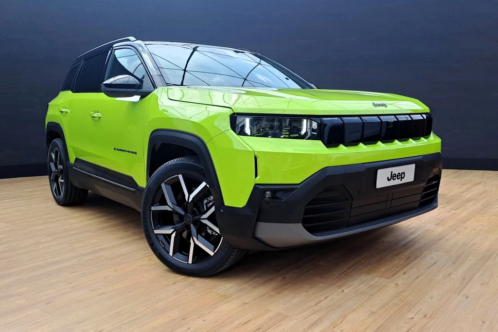 Jeep Compass będzie dostępny jako miękka hybryda, hybryda plug-in oraz elektryk. Nowoczesny samochód terenowy marki Jeep, w intensywnym zielonym kolorze, stojący w studiu na drewnianej podłodze. Samochód ma charakterystyczne reflektory LED, masywną kratkę wlotu powietrza oraz aluminiowe felgi. W tle ciemne ściany studia.