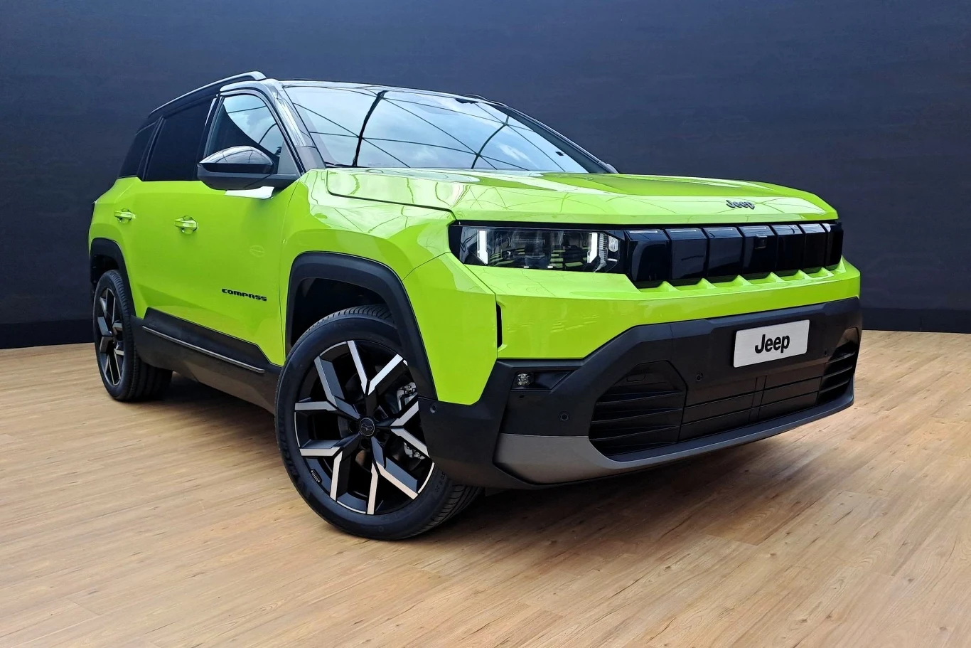 Nowoczesny samochód terenowy marki Jeep, w intensywnym zielonym kolorze, stojący w studiu na drewnianej podłodze. Samochód ma charakterystyczne reflektory LED, masywną kratkę wlotu powietrza oraz aluminiowe felgi. W tle ciemne ściany studia.