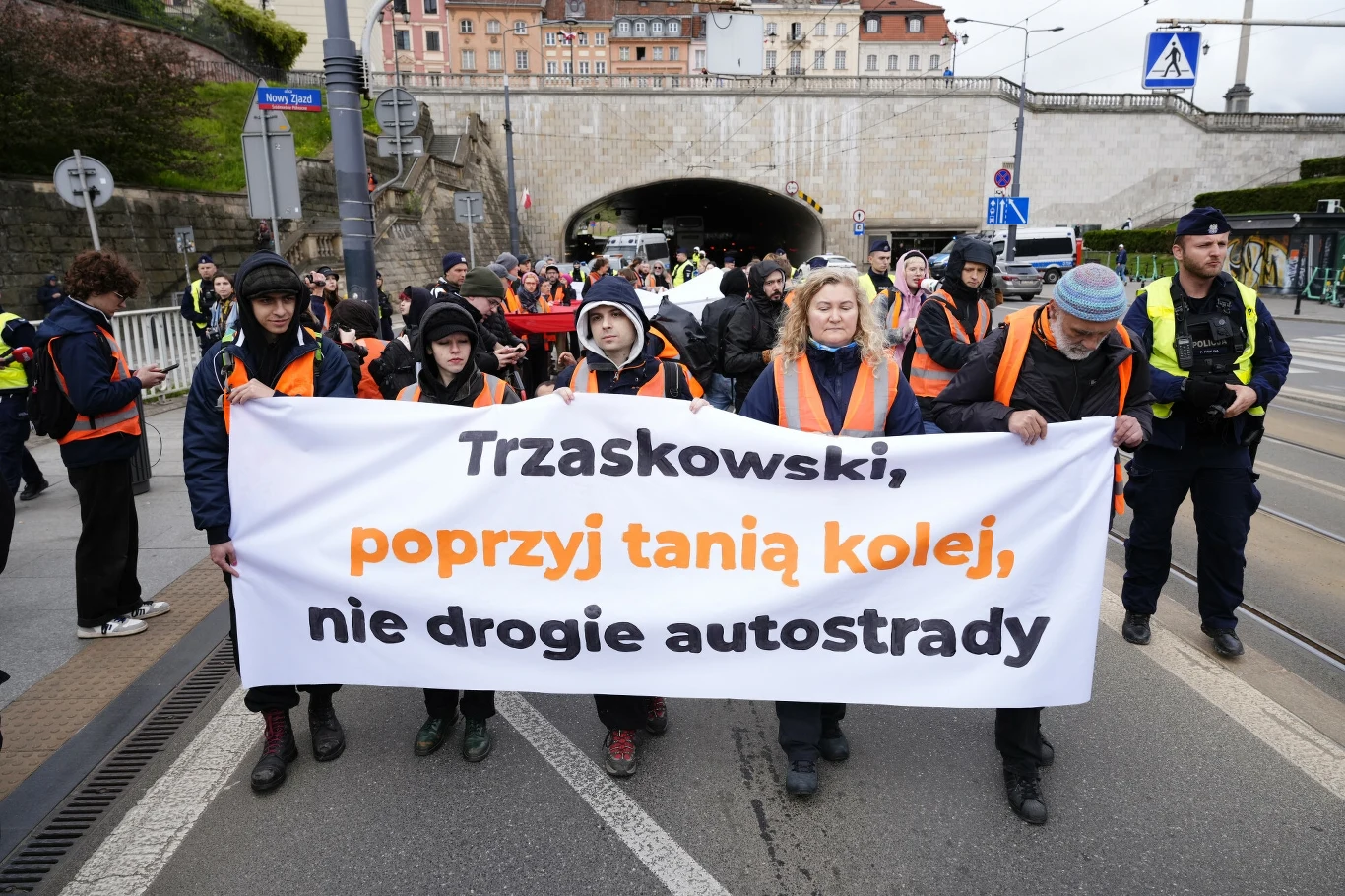 Ostatnie Pokolenie protestuje w Warszawie Grupa osób uczestniczących w proteście trzyma transparent z napisem apelującym do Trzaskowskiego o poparcie taniej kolei zamiast drogich autostrad. W tle widoczny most oraz osoby zabezpieczające wydarzenie.