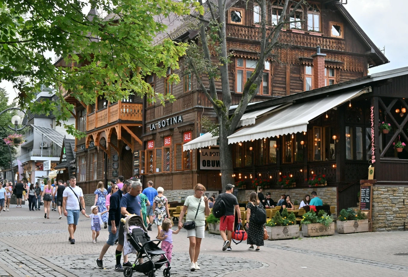 Ulica pełna spacerujących ludzi przed drewnianym budynkiem z napisem La Botina. Restauracja z zewnętrznym oświetleniem i kwiatami, otoczona zielonymi drzewami. Przechodnie z wózkiem dziecięcym i sklep obuwniczy obok.