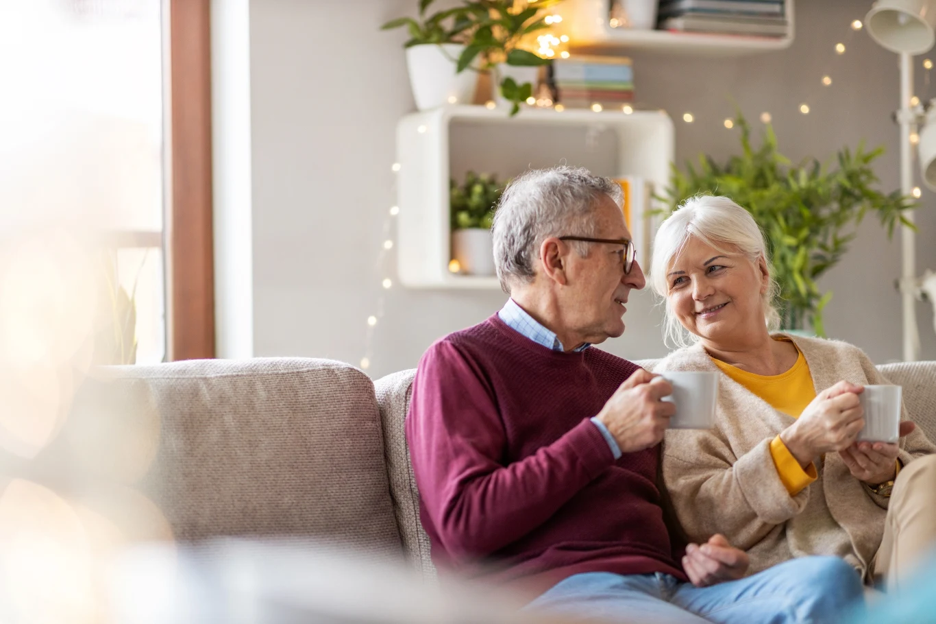 Starsza para siedząca na kanapie w przytulnym salonie, trzymająca filiżanki i rozmawiająca z uśmiechem. W tle widać rośliny doniczkowe i ozdobne światełka, które tworzą ciepłą atmosferę.