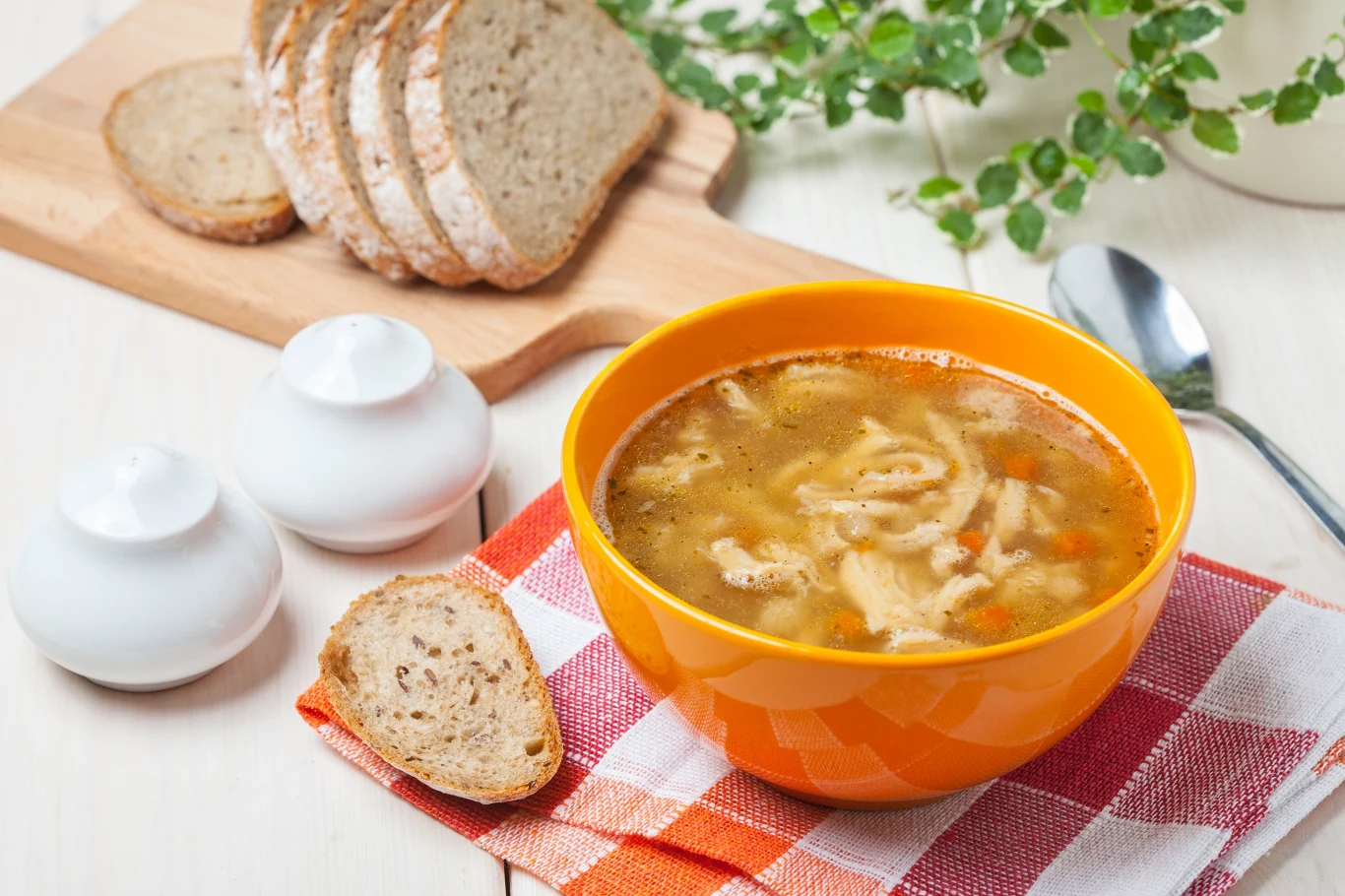 Pomarańczowa miska z zupą flaczki ustawiona na kolorowej serwetce obok chleba i przyprawników, w tle zielona roślina.