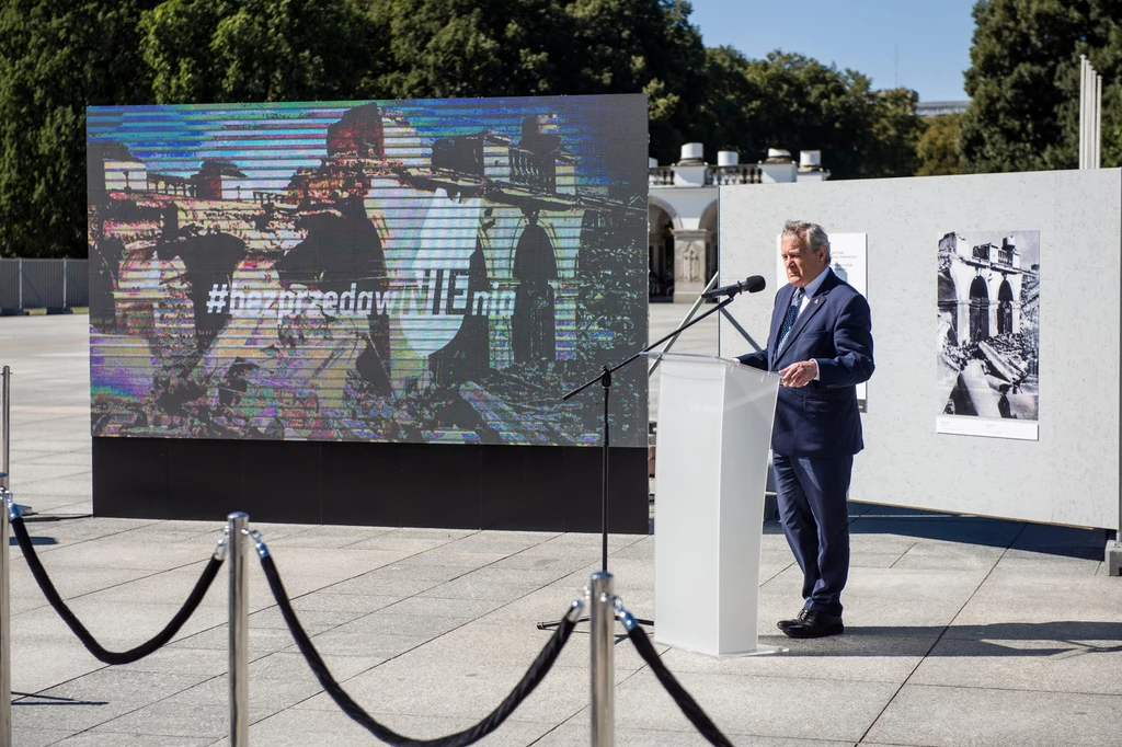 Otwarcie przez Piotra Glińskiego wystawy pod hasłem "Bez przedawnienia" na placu Piłsudskiego w Warszawie Mężczyzna w garniturze przemawia na otwartej przestrzeni, stojąc przy białej mównicy. W tle duży ekran wyświetlający obraz z napisem #naprzeddumniEMy, otoczenie jest spokojne, ludzie są nieobecni.