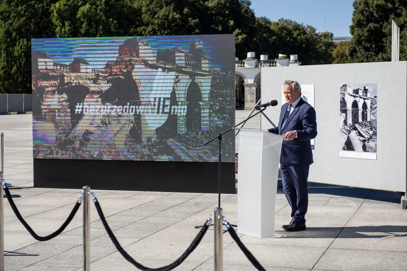 Mężczyzna w garniturze przemawia na otwartej przestrzeni, stojąc przy białej mównicy. W tle duży ekran wyświetlający obraz z napisem #naprzeddumniEMy, otoczenie jest spokojne, ludzie są nieobecni.