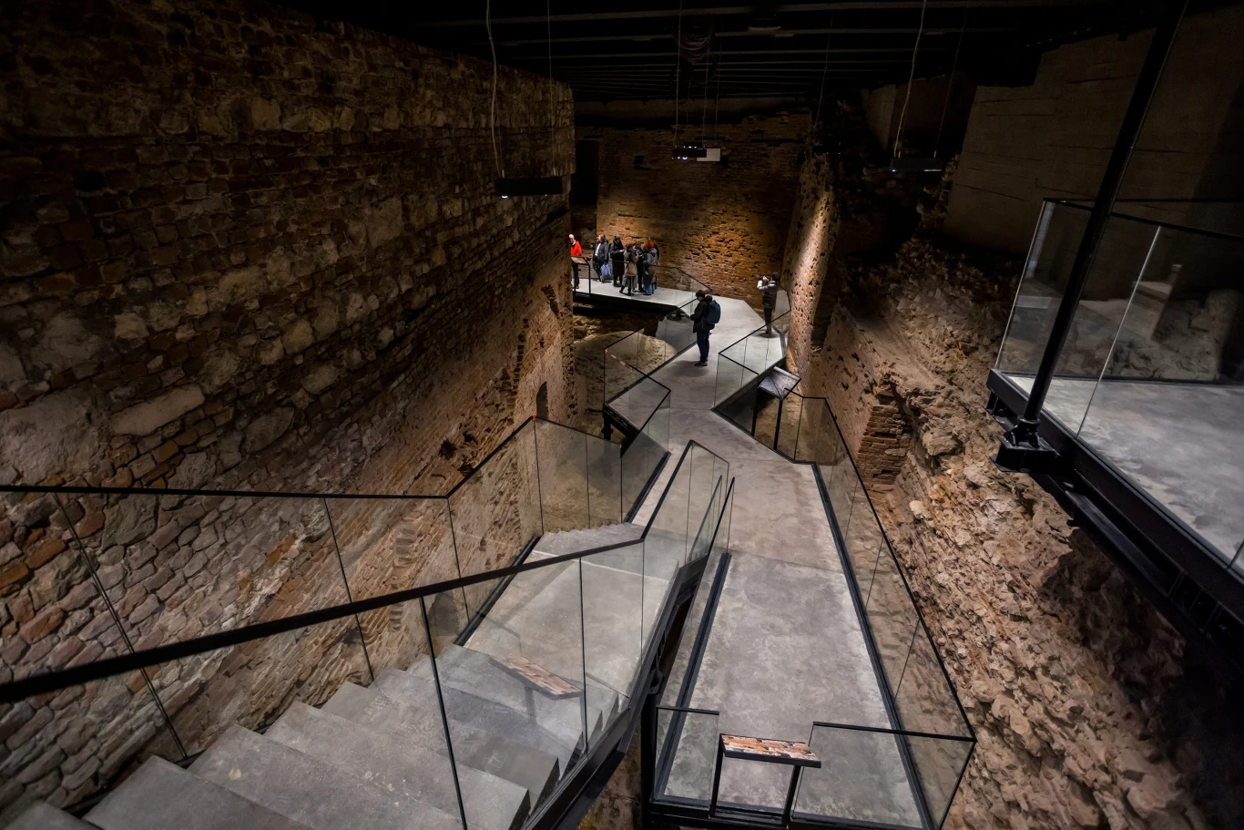 Nowa podziemna trasa na Wawelu zostanie otwarta już 23 maja 2025 roku. Oprócz wiekowych murów pełna będzie nowoczesnych urozmaiceń ukazujących historię miejsca. Widok na nowoczesne wnętrze ruin starego budynku z metalowymi i szklanymi balustradami. Strome schody prowadzą wzdłuż kamiennych ścian, tworząc futurystyczny kontrast do starożytnej architektury. Grupa ludzi na końcu korytarza.