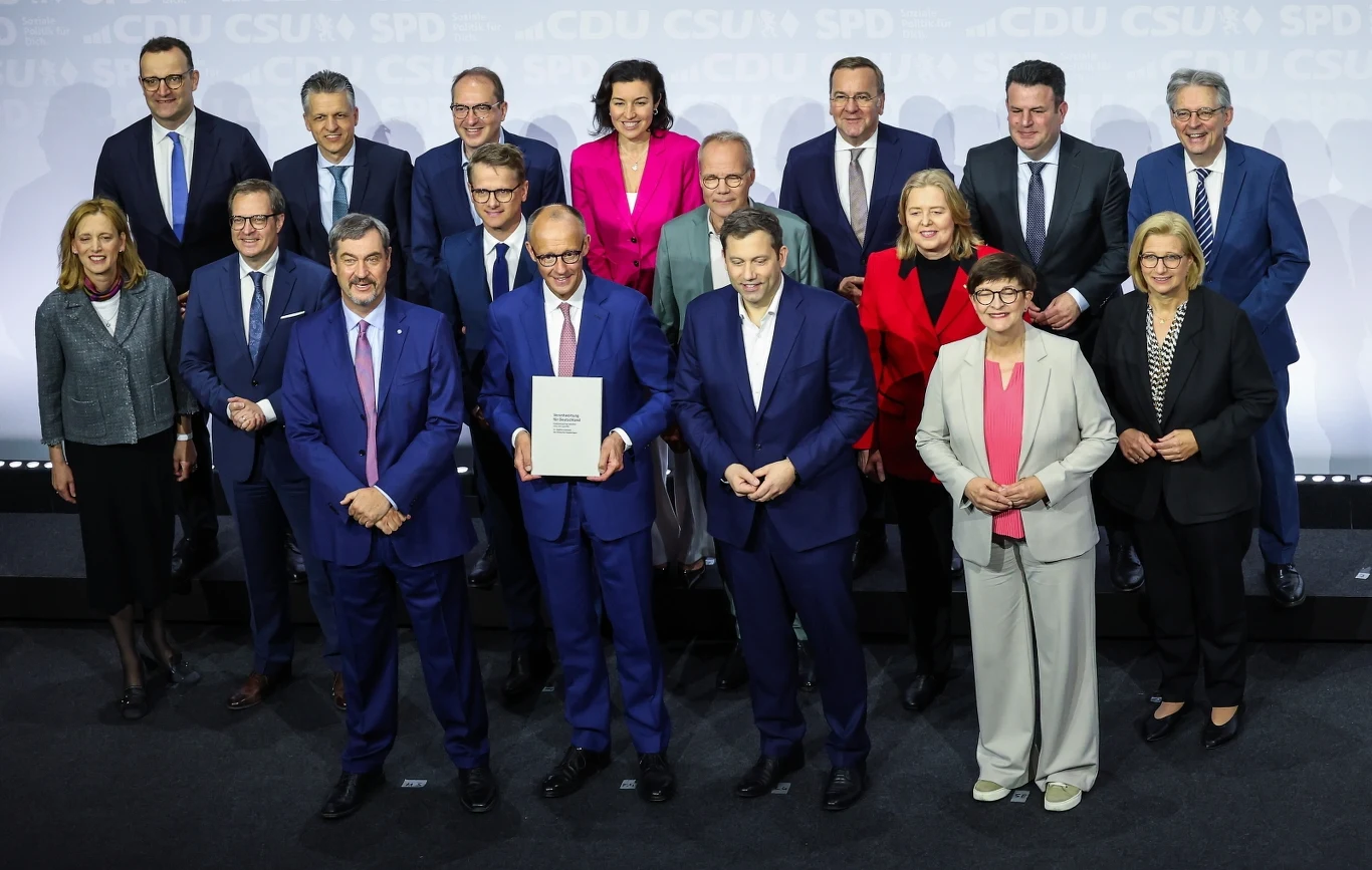 Chadecy i socjaldemokraci podpisali umowę koalicyjną Grupa osób w formalnym stroju pozuje do wspólnego zdjęcia. Na pierwszy planie znajduje się mężczyzna w niebieskim garniturze trzymający teczkę. W tle widać kilka osób w eleganckich marynarkach i kostiumach, stojących na podwyższeniu.