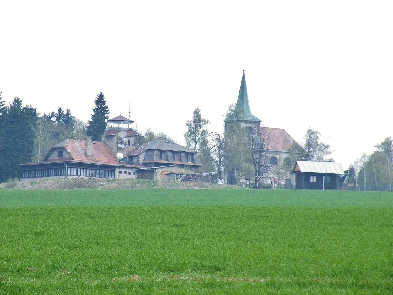 Wiejski krajobraz z zieloną łąką na pierwszym planie i zespołem budynków w oddali, w tym wieżą kościoła z zielonym dachem. Budynki otoczone drzewami, co nadaje malowniczy i spokojny wygląd.