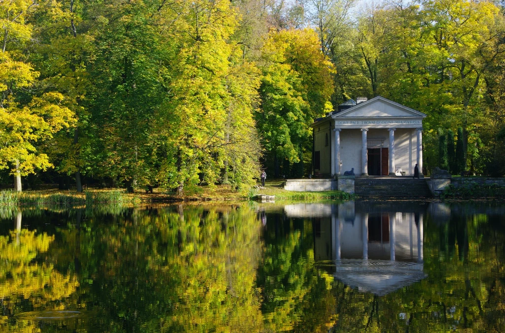 Park o każdej porze roku prezentuje się urokliwie. Na zdjęciu Świątynia Diany Budynek nad stawem otoczony bujną zielenią drzew odbija się w spokojnej tafli wody. Architektura przypomina klasyczny styl. Kolory jesieni dominują w krajobrazie, tworząc harmonijną i spokojną scenerię.