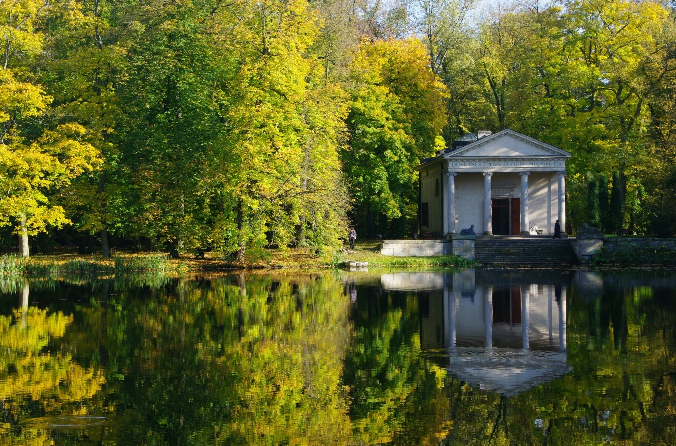 Budynek nad stawem otoczony bujną zielenią drzew odbija się w spokojnej tafli wody. Architektura przypomina klasyczny styl. Kolory jesieni dominują w krajobrazie, tworząc harmonijną i spokojną scenerię.