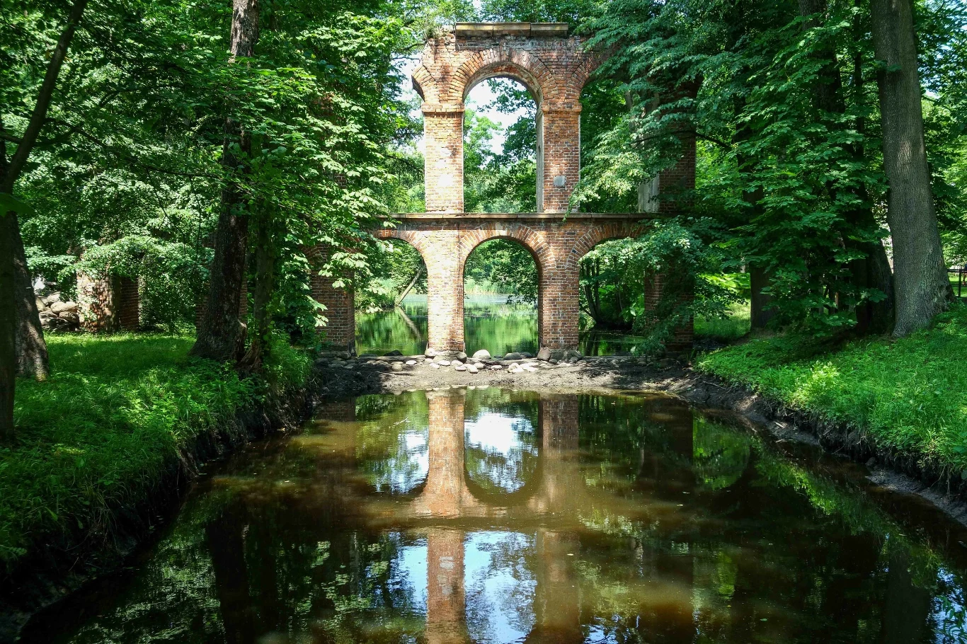 Zabytkowy most ceglany w parku otoczonym drzewami, odbijający się w spokojnej wodzie kanału, tworząc malowniczy krajobraz naturalny.