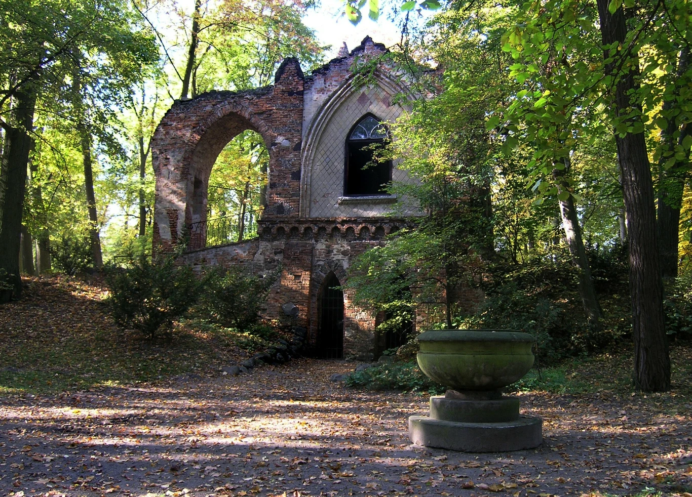 Ruiny gotyckiego budynku otoczone zielenią, z wysokim łukowatym oknem i ceglanymi ścianami. W centrum placu znajduje się kamienna misa, a otoczenie pełne jest drzew i liści.”