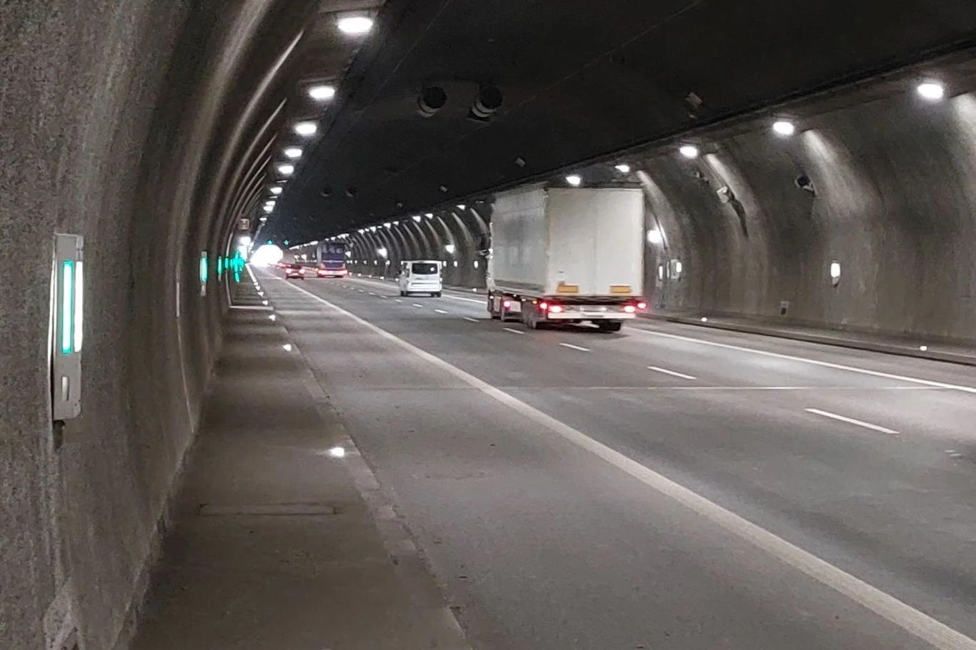 Samochody poruszające się w tunelu autostradowym, widoczny pojazd ciężarowy oraz samochód osobowy jadące w tym samym kierunku. Oświetlenie tunelu tworzy regularny wzór na suficie i ścianach, a w oddali widać wylot tunelu.