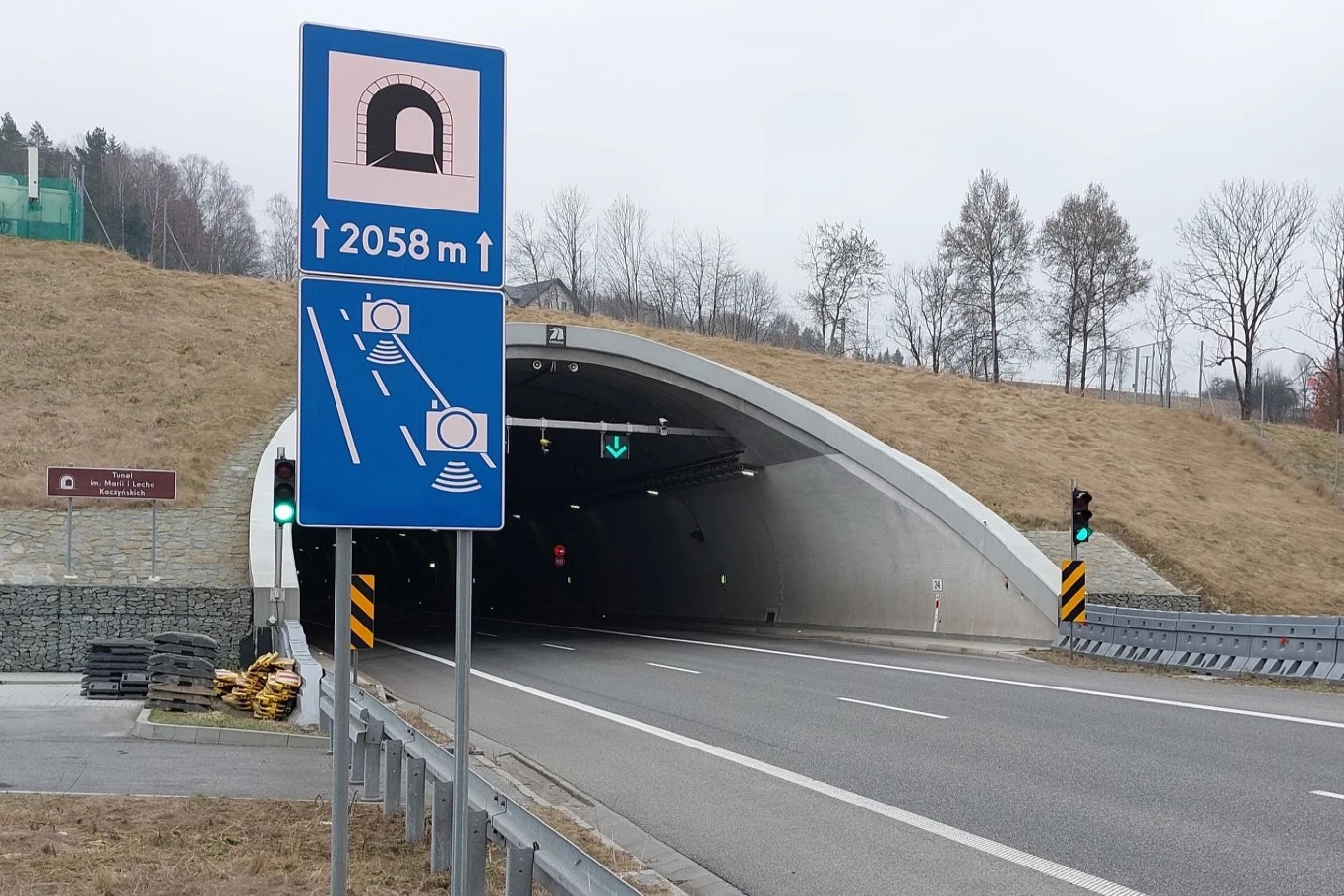 Tunel drogowy z dobrze widocznymi znakami drogowymi i sygnalizacją świetlną. Na pierwszym planie znak informacyjny ze strzałkami kierującymi do tunelu o długości 2058 m.