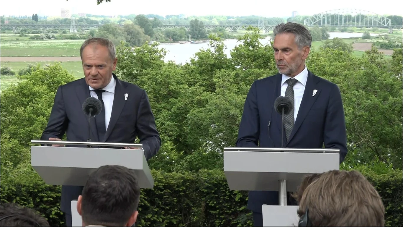 Donald Tusk Dwóch mężczyzn w garniturach stojących przy mównicach w otoczeniu zieleni, z widokiem na most i rzekę w tle.