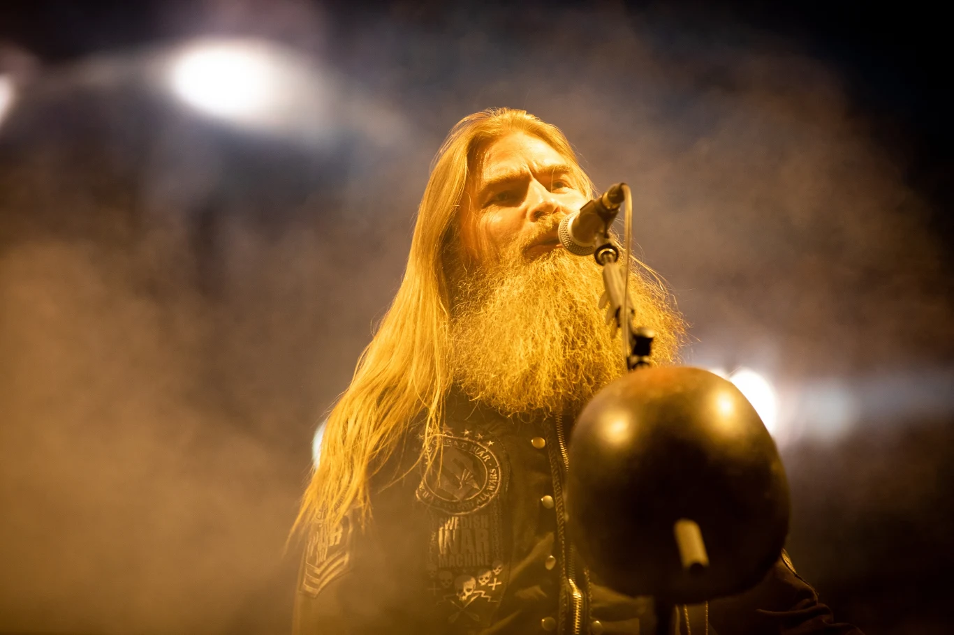 Pär Sundström jest basistą grupy Sabaton Muzyk o długiej brodzie śpiewa do mikrofonu na scenie, w tle widoczne są intensywne światła reflektorów, atmosfera jest pełna dymu i tajemniczości.