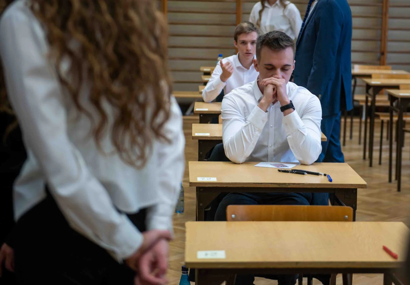 Matura 2025. Polonistka o arkuszu: przyjazny, ale nie należał do banalnych Sala egzaminacyjna z osobami w jasnych koszulach siedzącymi przy oddzielnych stolikach, na pierwszym planie młody mężczyzna skoncentrowany nad arkuszem, w tle osoba pokazująca gest kciuka w górę.