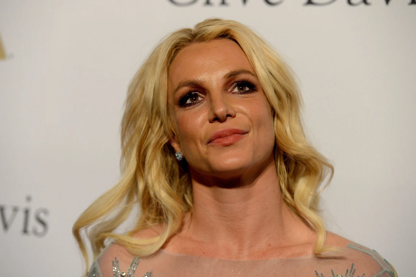 Britney Spears Kobieta o blond włosach i delikatnym uśmiechu, ubrana w elegancką sukienkę, stoi na tle białej ściany z logo Clive Davis widocznym w tle. Ma wyrazisty makijaż podkreślający oczy.