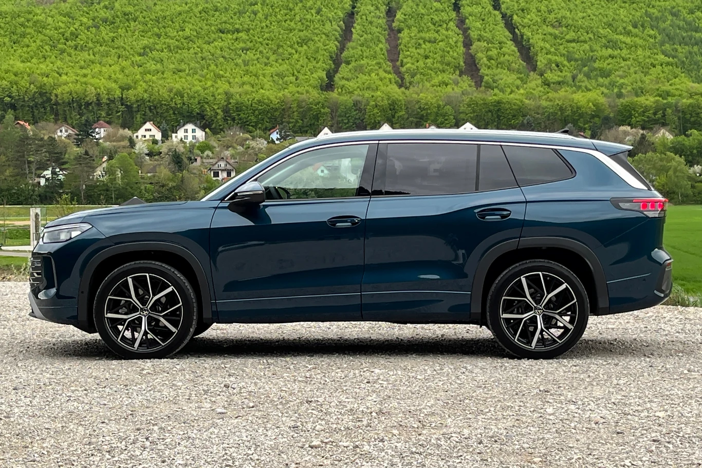 Volkswagen Tayron wchodzi do oferty, jako siedmiomiejscowy następca modelu Tiguan Allspace. Nowoczesny samochód typu SUV w kolorze granatowym stoi zaparkowany na żwirowym podłożu. W tle widoczne są zielone wzgórza oraz rząd białych domów, co tworzy spokojny krajobraz.