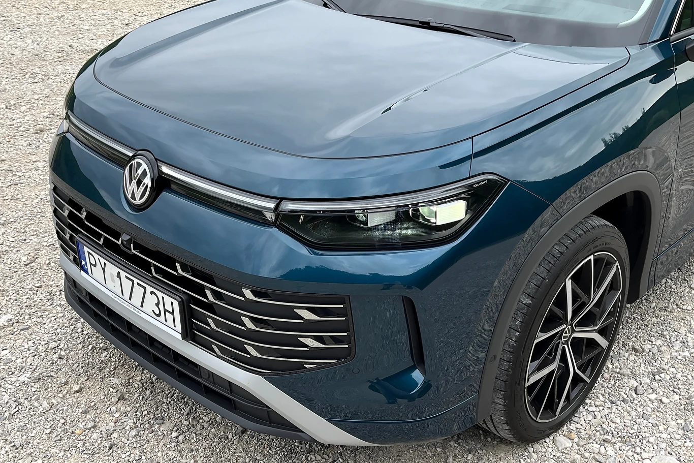 Volkswagen Tayron to kwintesencja niemieckiej motoryzacji. Część przednia ciemnoniebieskiego samochodu marki Volkswagen na żwirowym podłożu. Widoczne reflektory, zderzak oraz charakterystyczne logo marki na masce pojazdu. Nowoczesna stylistyka i elegancki wygląd.