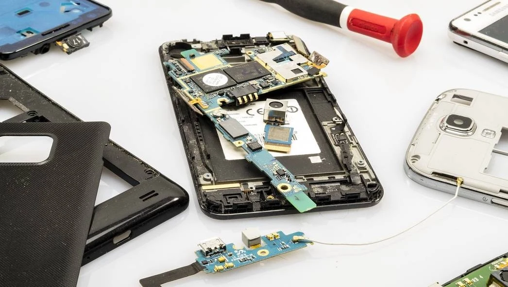 Rozłożony telefon komórkowy z widocznymi komponentami takimi jak płyta główna i obudowa, otoczony przez narzędzie i inne części, co sugeruje proces serwisowy lub naprawczy.